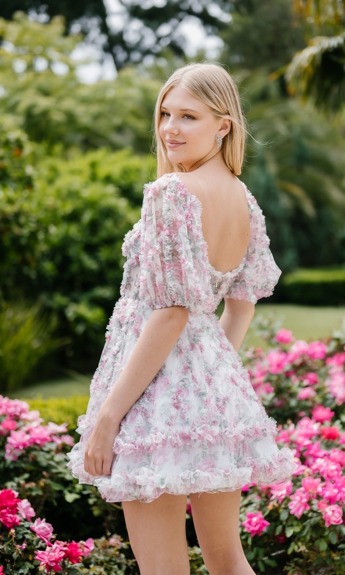 A woman with long blonde hair stands in a garden surrounded by pink flowers, wearing the Aleta 1304 short sleeve short print prom dress.
